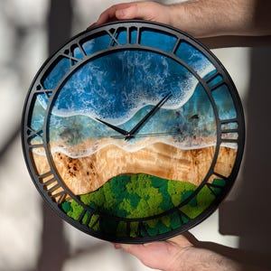 Pu&ograve; includere: Orologio da parete rotondo con cornice nera e numeri romani. Il quadrante presenta una scena di spiaggia con onde blu dell'oceano, una riva sabbiosa e una collina verde e muschiosa. Le lancette dell'orologio sono nere.