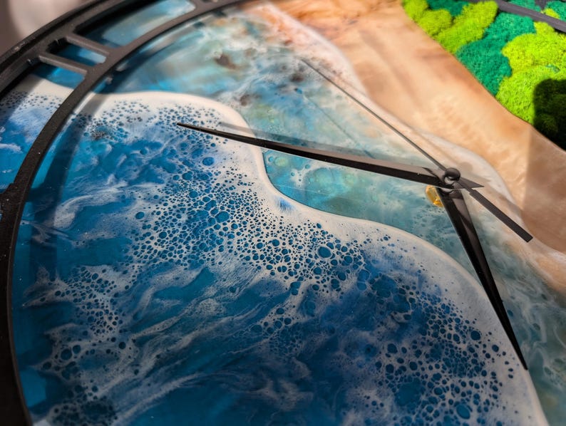 Pu&ograve; includere: Primo piano di un orologio da parete con un design in resina raffigurante onde oceaniche. Il quadrante dell'orologio presenta sfumature di blu e bianco, con lancette nere e una cornice nera. Il design assomiglia a una scena di spiaggia.