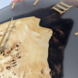 May include: A round wall clock with a natural wood grain face, dark gray resin, and gold-colored Roman numerals and hands. The clock's face has a unique, organic pattern, and the hands point to the time.