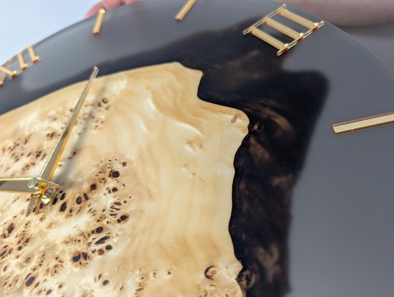 May include: A round wall clock with a natural wood grain face, dark gray resin, and gold-colored Roman numerals and hands. The clock's face has a unique, organic pattern, and the hands point to the time.