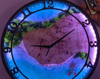 May include: A black circular clock with a resin beach scene inside. The clock face has a blue and green ocean with a sandy beach and a small island with green moss. The clock has a black frame with white numbers and a black hand. The clock is lit up with a blue light.