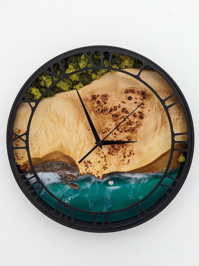 May include: A round wall clock with a black frame and Roman numerals. The clock face features a natural wood grain design with a turquoise resin ocean and small boats. Green moss and a wooden landscape are also visible.