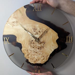 May include: A round wall clock with a light wood grain center and a dark gray resin border. The clock features gold Roman numerals and hands. The clock is being held by two hands.