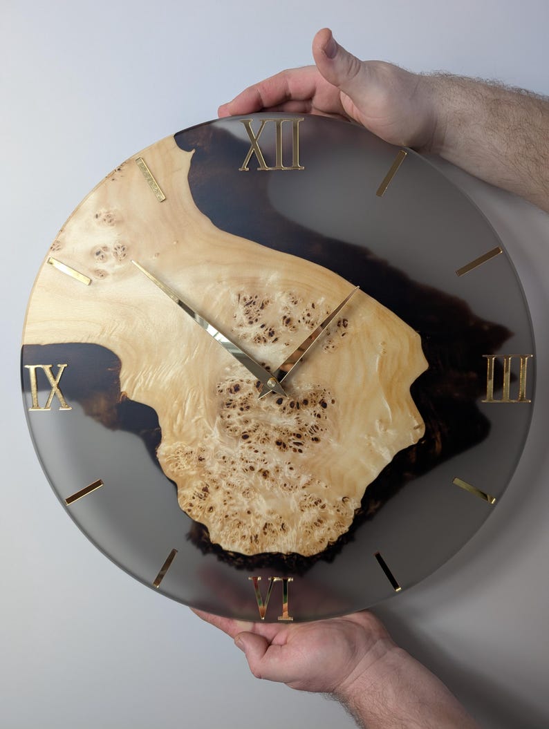 May include: A round wall clock with a light wood grain center and a dark gray resin border. The clock features gold Roman numerals and hands. The clock is being held by two hands.