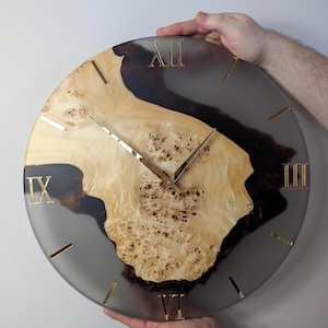 May include: A round wall clock with a natural wood and resin design. The clock face features a light-colored wood center with dark resin accents and gold-colored Roman numerals and hands. The clock is being held by two hands.