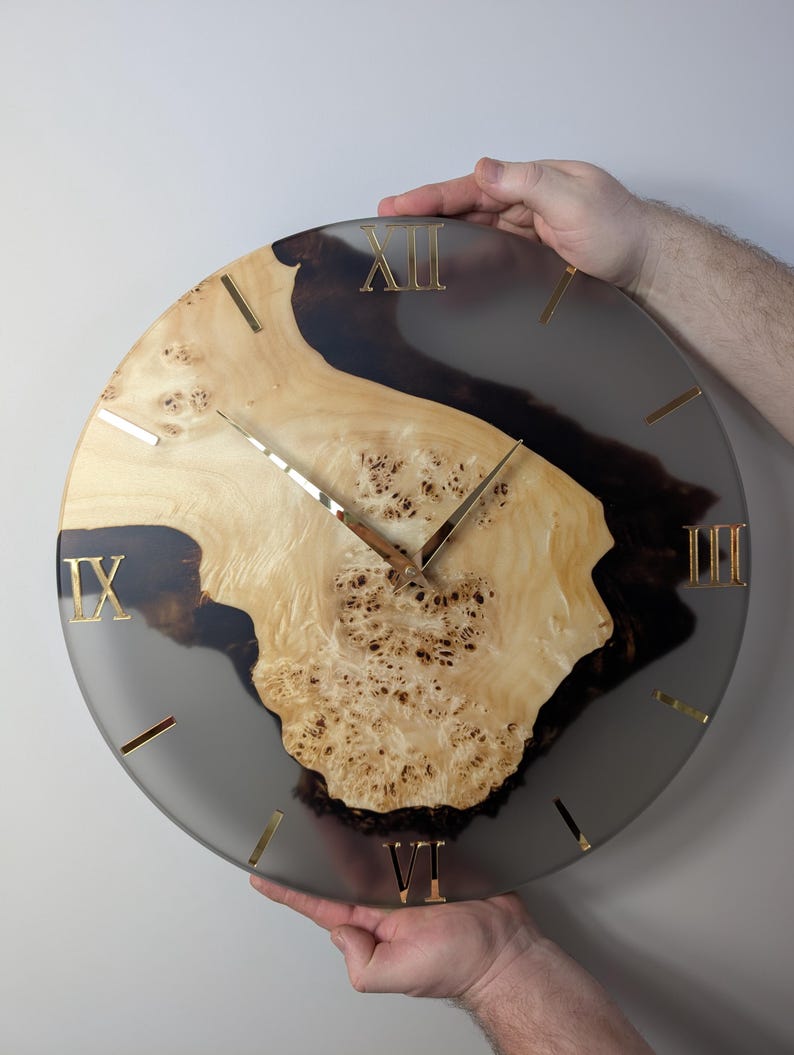 May include: A round wall clock with a natural wood and resin design. The clock face features a light-colored wood center with dark resin accents and gold-colored Roman numerals and hands. The clock is being held by two hands.
