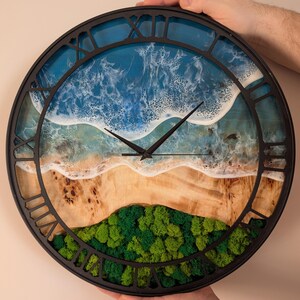 Pu&ograve; includere: Un orologio da parete rotondo con cornice nera e numeri romani. Il quadrante presenta una scena di spiaggia con oceano blu, onde bianche, una riva sabbiosa e muschio verde. L'orologio &egrave; tenuto da due mani.