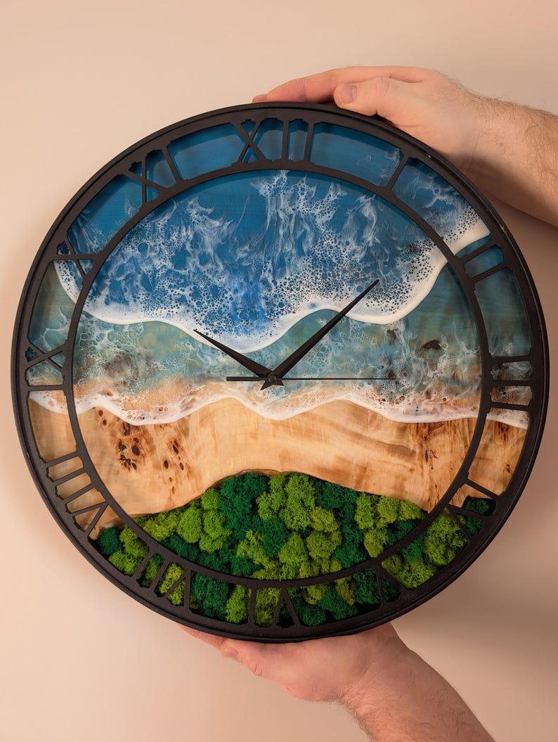 Pu&ograve; includere: Un orologio da parete rotondo con cornice nera e numeri romani. Il quadrante presenta una scena di spiaggia con oceano blu, onde bianche, una riva sabbiosa e muschio verde. L'orologio &egrave; tenuto da due mani.