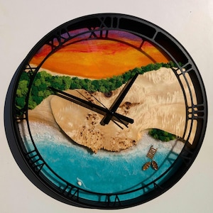 May include: A round clock with a black frame and Roman numeral markings. The clock face features a resin art design of a beach scene with a sunset, sand, and ocean waves.