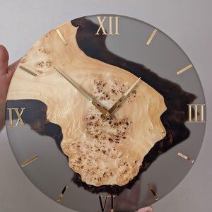 May include: A round wall clock with a natural wood burl center and a gray resin border. Gold Roman numerals mark the hours. The clock hands are gold. The wood has a light brown color with dark brown accents.