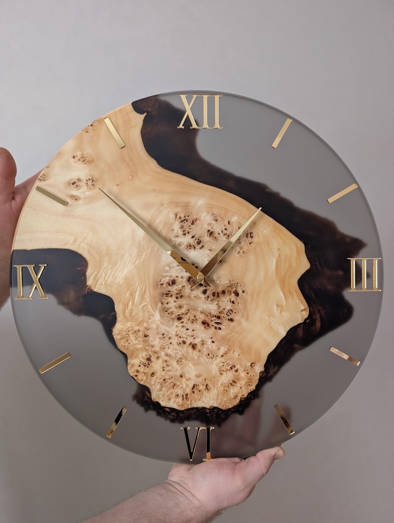 May include: A round wall clock with a natural wood burl center and a gray resin border. Gold Roman numerals mark the hours. The clock hands are gold. The wood has a light brown color with dark brown accents.