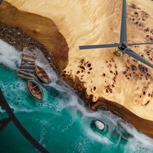 May include: A unique wall clock featuring a beach scene with a wooden dock and two small boats. The clock face is made of wood and resin, with black clock hands and a black metal frame. The clock measures approximately 12 inches in diameter.