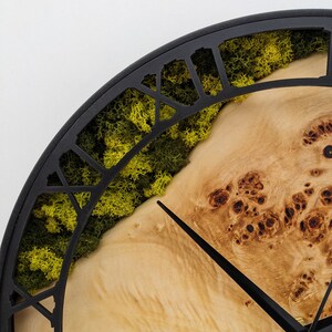 May include: A close-up of a round wall clock with a black metal frame. The clock face features a light-colored wood grain design, with a section of green moss. Black clock hands point to the time.