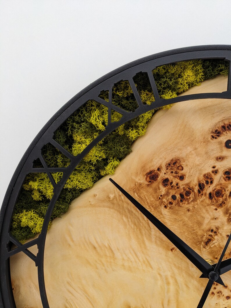 May include: A close-up of a round wall clock with a black metal frame. The clock face features a light-colored wood grain design, with a section of green moss. Black clock hands point to the time.