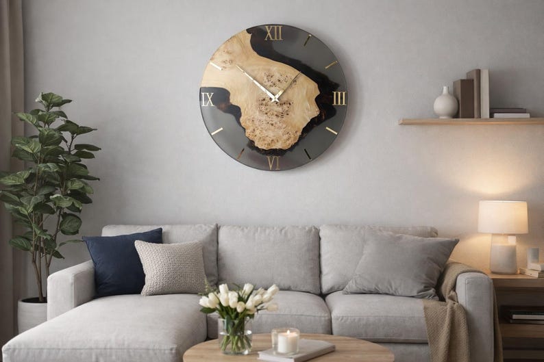 May include: A large, round wall clock with a natural wood center and a dark gray epoxy resin border. The clock features gold Roman numerals and hands. The clock is mounted on a light gray wall above a gray sofa.