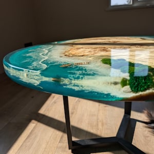 May include: A round coffee table with a unique epoxy resin top featuring a blue ocean scene with a small island and green foliage. The table has a black metal base with a geometric design.