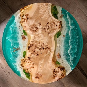 Puede incluir: Mesa redonda de madera con diseño de escena de playa. La mesa presenta un centro de veta de madera marrón claro, rodeado por un océano de resina turquesa. Tortugas en miniatura, estrellas de mar y sillas de playa están incrustadas en la resina.