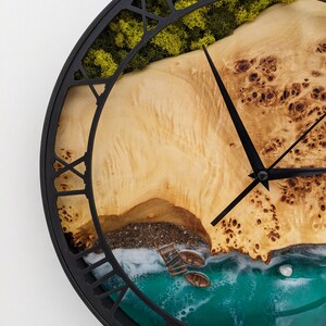 May include: A round wall clock with a black metal frame and Roman numeral hour markers. The clock face features a detailed beach scene with a wooden shore, green moss, and turquoise water. Two black clock hands indicate the time.