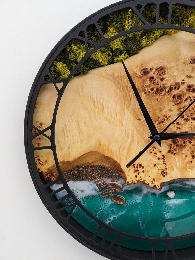 May include: A round wall clock with a black metal frame and Roman numeral hour markers. The clock face features a detailed beach scene with a wooden shore, green moss, and turquoise water. Two black clock hands indicate the time.