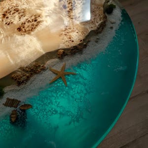 May include: A round, teal resin table top with a white sandy beach and a small island made of wood. The island has a brown, natural wood grain and a starfish on the beach.