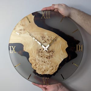 May include: A round wall clock with a light wood and dark wood grain design. The clock face is a light gray color with gold Roman numerals and gold clock hands. The clock is approximately 12 inches in diameter.