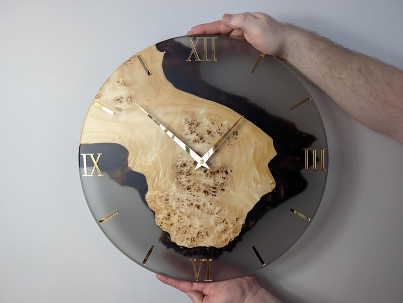 May include: A round wall clock with a light wood and dark wood grain design. The clock face is a light gray color with gold Roman numerals and gold clock hands. The clock is approximately 12 inches in diameter.