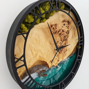 May include: A round wall clock with a black frame and Roman numeral hour markers. The clock face features a beach scene with a wooden surface, green moss, and a turquoise ocean. The clock hands are black.