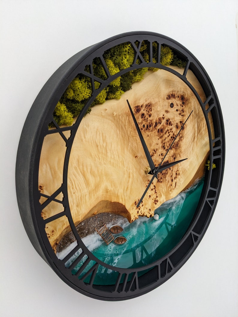 May include: A round wall clock with a black frame and Roman numeral hour markers. The clock face features a beach scene with a wooden surface, green moss, and a turquoise ocean. The clock hands are black.