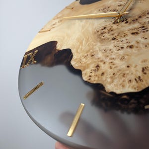 May include: Close-up of a round wall clock with a unique design. The clock face features a combination of light wood and gray resin. Gold-colored Roman numerals and hands complete the design. The clock's diameter is approximately 12 inches.