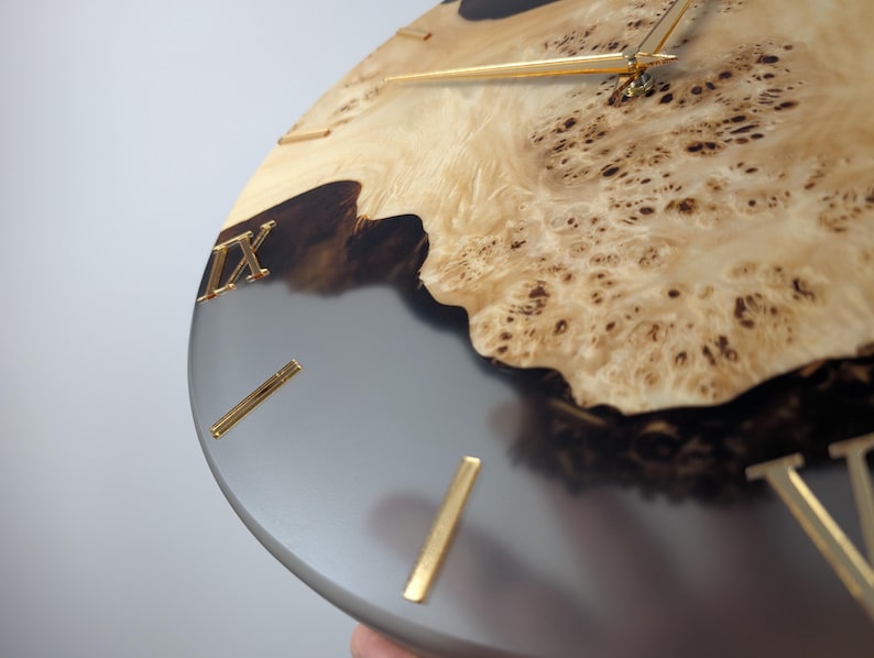 May include: Close-up of a round wall clock with a unique design. The clock face features a combination of light wood and gray resin. Gold-colored Roman numerals and hands complete the design. The clock's diameter is approximately 12 inches.
