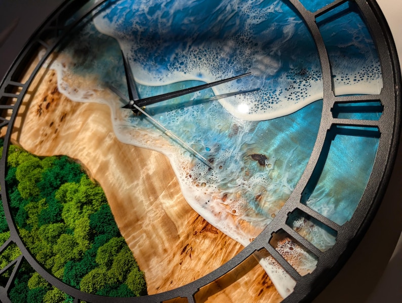 Pu&ograve; includere: Un orologio da parete rotondo con un design a scena di spiaggia. Il quadrante dell'orologio presenta una rappresentazione dettagliata di una spiaggia con onde blu dell'oceano, rive sabbiose e fogliame verde. L'orologio ha lancette nere e una cornice nera.