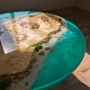 May include: A round, turquoise and white resin table top with a beach scene. The table top features a sandy beach with brown rocks, a starfish, and a few seashells. The beach is surrounded by turquoise water.