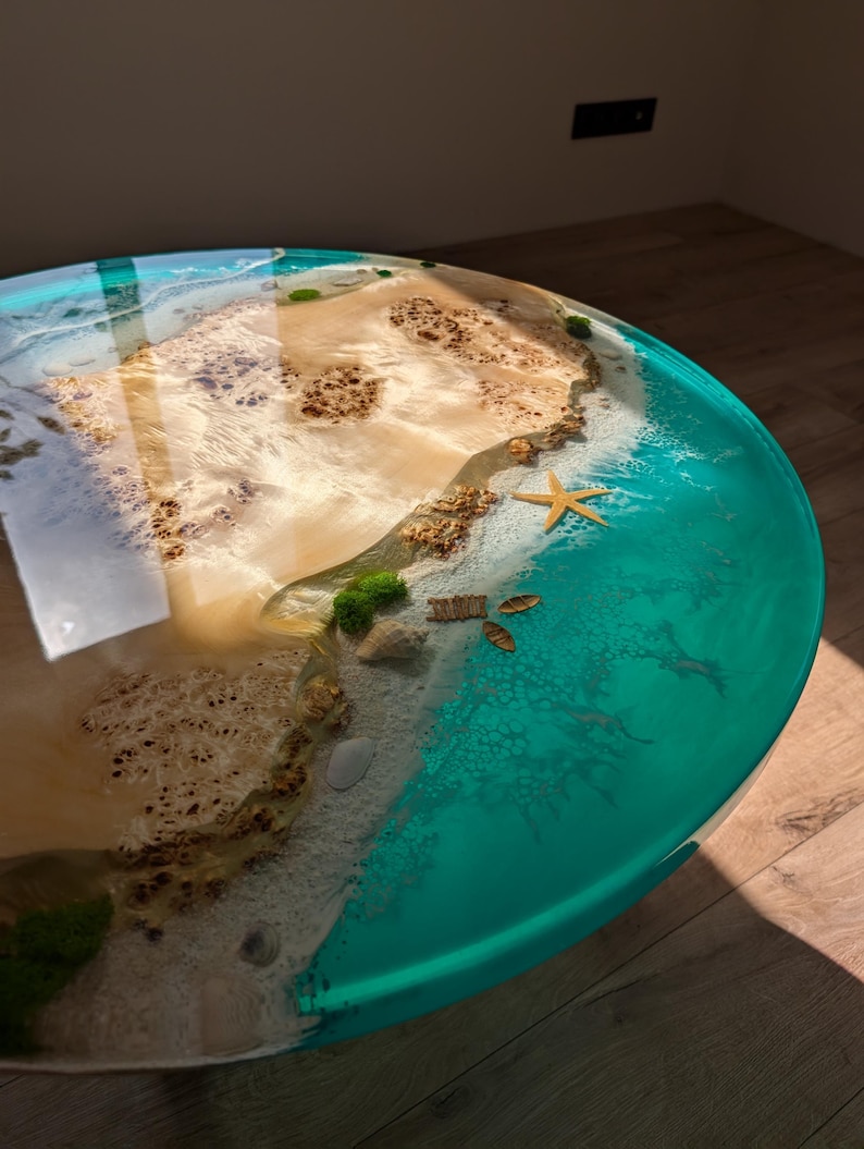May include: A round, turquoise and white resin table top with a beach scene. The table top features a sandy beach with brown rocks, a starfish, and a few seashells. The beach is surrounded by turquoise water.