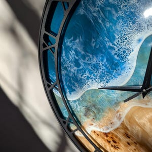 Pu&ograve; includere: Un orologio da parete rotondo con una cornice in metallo nero. Il quadrante dell'orologio presenta una scena oceanica dettagliata con onde blu e bianche, una spiaggia sabbiosa e fogliame verde. Le lancette dell'orologio sono nere.