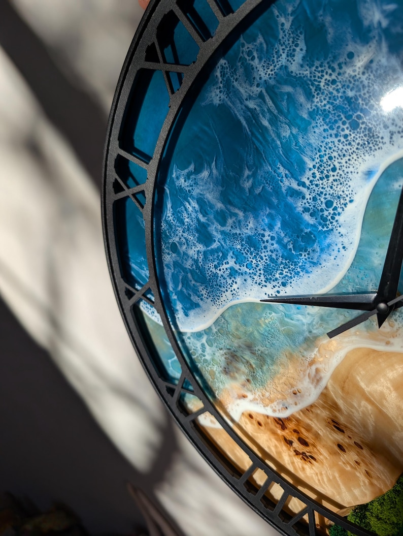 Pu&ograve; includere: Un orologio da parete rotondo con una cornice in metallo nero. Il quadrante dell'orologio presenta una scena oceanica dettagliata con onde blu e bianche, una spiaggia sabbiosa e fogliame verde. Le lancette dell'orologio sono nere.