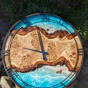 Puede incluir: Un reloj redondo con marco negro y números romanos. La esfera del reloj presenta un diseño de veta de madera con un diseño de ola de resina epoxi azul.