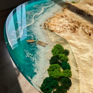 May include: A round, turquoise resin table top with a miniature beach scene. The scene includes a sandy beach, a small boat, a wooden dock, and green moss trees.