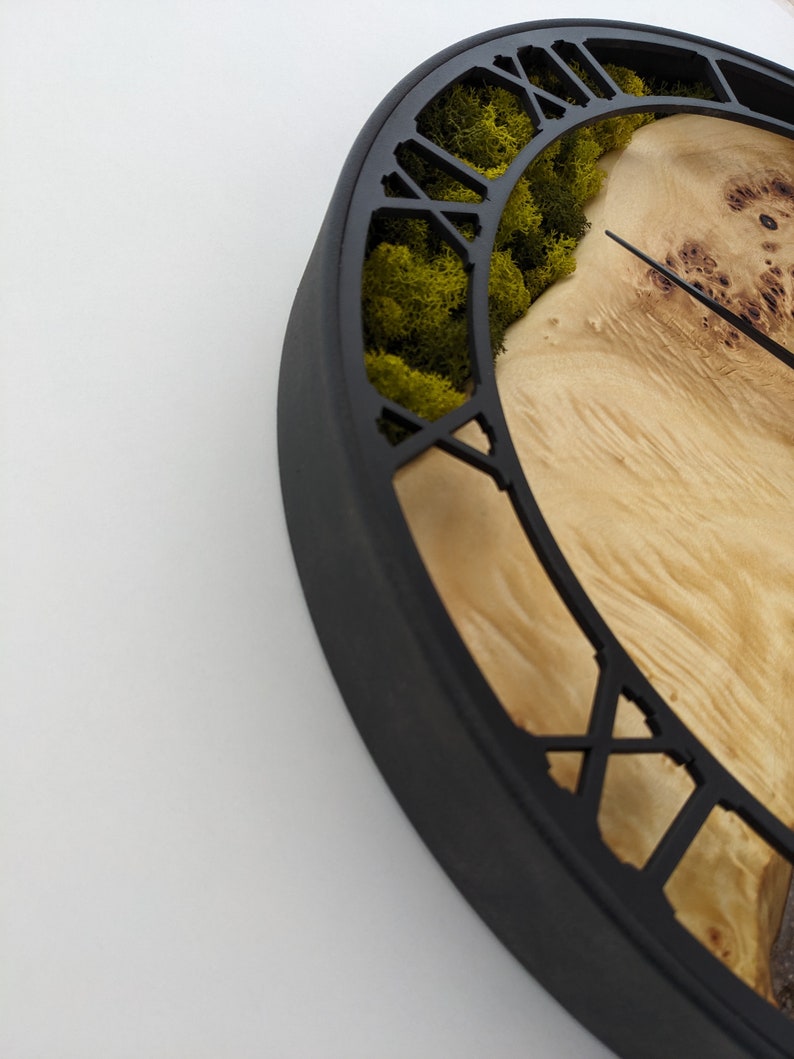May include: A unique wall clock with a black frame and Roman numerals. The clock face features natural wood grain and a section of green moss. The clock hands are black.
