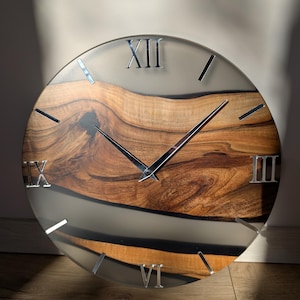 Könnte beinhalten: Eine runde Wanduhr mit einem Holzkern und einem transparenten Harzrand. Der Holzteil hat ein natürliches Maserungsmuster. Silberne römische Ziffern markieren die Stunden. Die Uhrzeiger sind silberfarben.