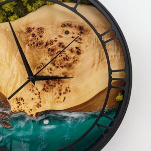 May include: A round wall clock with a black metal frame and Roman numeral hour markers. The clock face features a light-colored wood grain design with a turquoise resin ocean scene and green moss accents.