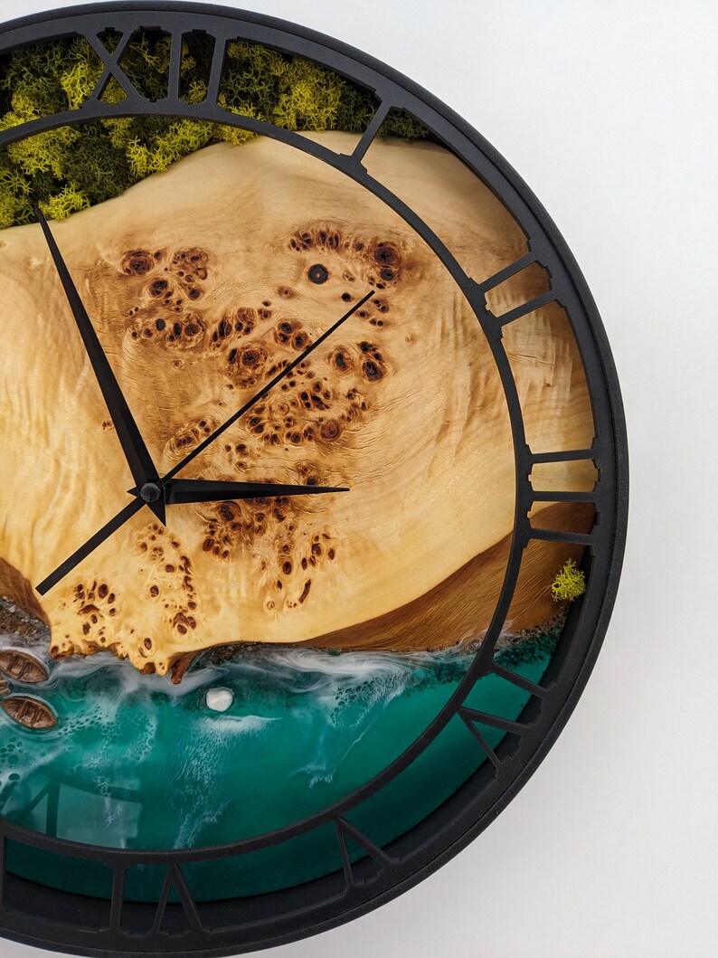 May include: A round wall clock with a black metal frame and Roman numeral hour markers. The clock face features a light-colored wood grain design with a turquoise resin ocean scene and green moss accents.