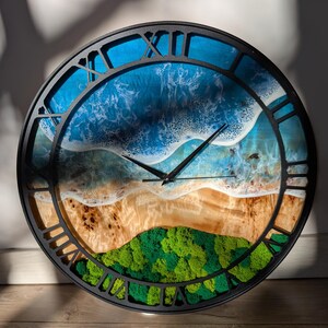 Pu&ograve; includere: Un orologio da parete rotondo con cornice nera e numeri romani. Il quadrante presenta una scena di spiaggia con onde blu dell'oceano, una riva sabbiosa e colline verdi e muschiose. Le lancette dell'orologio sono nere.