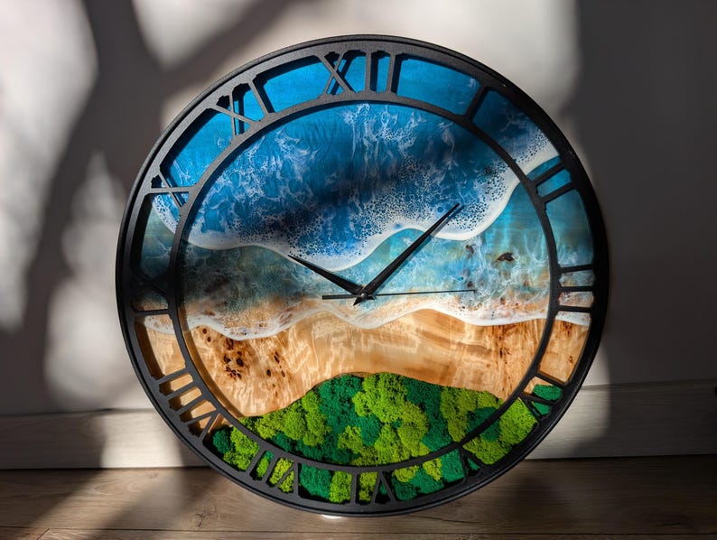 Pu&ograve; includere: Un orologio da parete rotondo con cornice nera e numeri romani. Il quadrante presenta una scena di spiaggia con onde blu dell'oceano, una riva sabbiosa e colline verdi e muschiose. Le lancette dell'orologio sono nere.