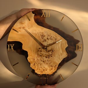 May include: A round wall clock with a light-colored wood grain center and gold-colored Roman numerals. The clock hands are gold, and the clock is held by a person's hands.