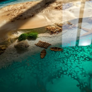 May include: A close-up of a resin art piece depicting a beach scene with turquoise water, white sand, and small wooden boats. The water has a bubbly texture and the sand has a light brown color.