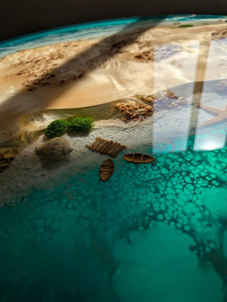 May include: A close-up of a resin art piece depicting a beach scene with turquoise water, white sand, and small wooden boats. The water has a bubbly texture and the sand has a light brown color.