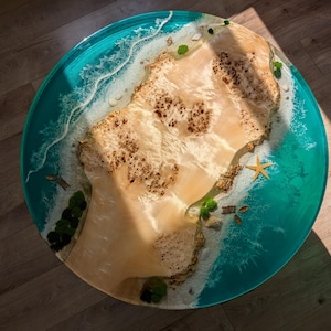 May include: A round turquoise resin table top with a white wave design and a beige island in the center. The island has a brown wood grain pattern and a small starfish on the beach.