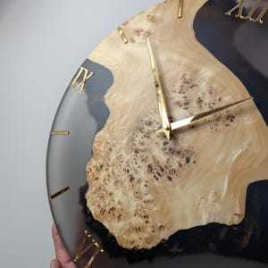 May include: A round wall clock with a light wood grain center and a dark gray resin border. Gold-colored Roman numerals mark the hours. The clock hands are also gold. The clock is being held by a person.