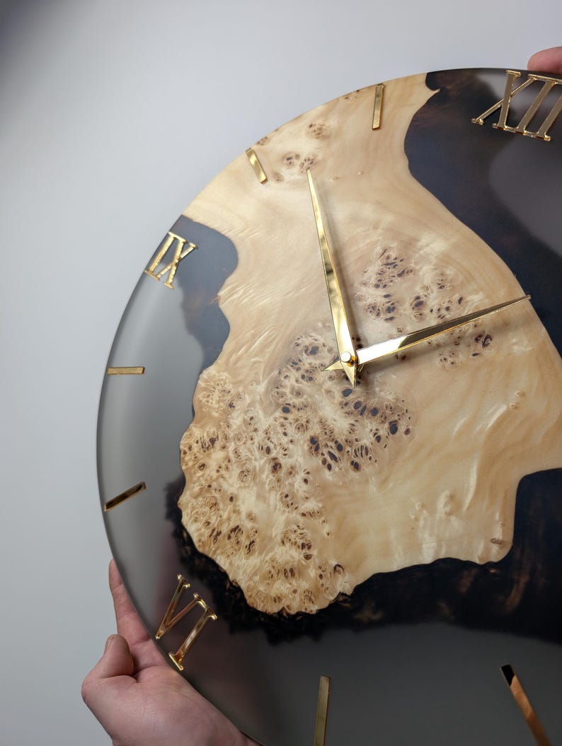 May include: A round wall clock with a light wood grain center and a dark gray resin border. Gold-colored Roman numerals mark the hours. The clock hands are also gold. The clock is being held by a person.