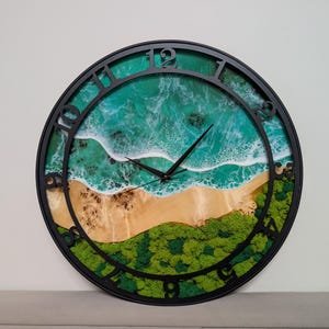 Peut inclure: Une horloge murale ronde avec un cadre noir. Le cadran de l'horloge représente une scène de plage avec de l'eau turquoise, des vagues blanches, une plage de sable et du feuillage vert. Les aiguilles de l'horloge sont noires.